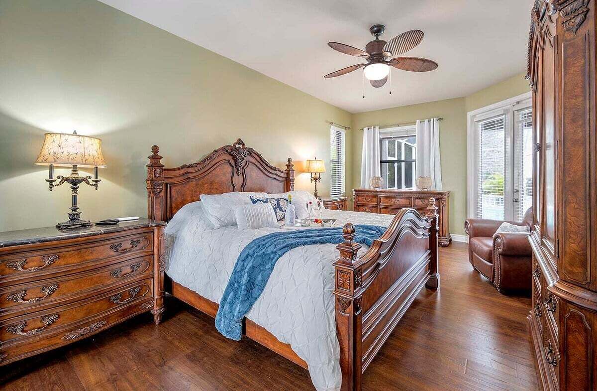 A bedroom with a large wooden bed, two bedside tables with lamps, a ceiling fan, a brown leather chair, and a wooden wardrobe.