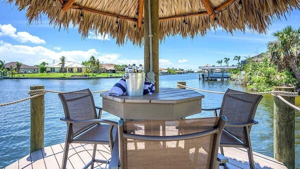 A wooden deck with a thatched roof umbrella, two chairs, and a table with a bucket and towel. - Surfside - Bikes and Weight Bench (Cape Coral)
