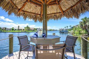 A wooden deck with a thatched roof umbrella, two chairs, and a table with a bucket and towel.