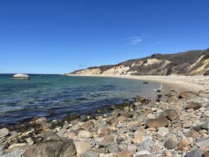 A rocky beach with clear water, a sandy shore, and a hilly landscape in the background. - Oak Bluffs 4BR • 5 Min Drive to Downtown (Oak Bluffs)