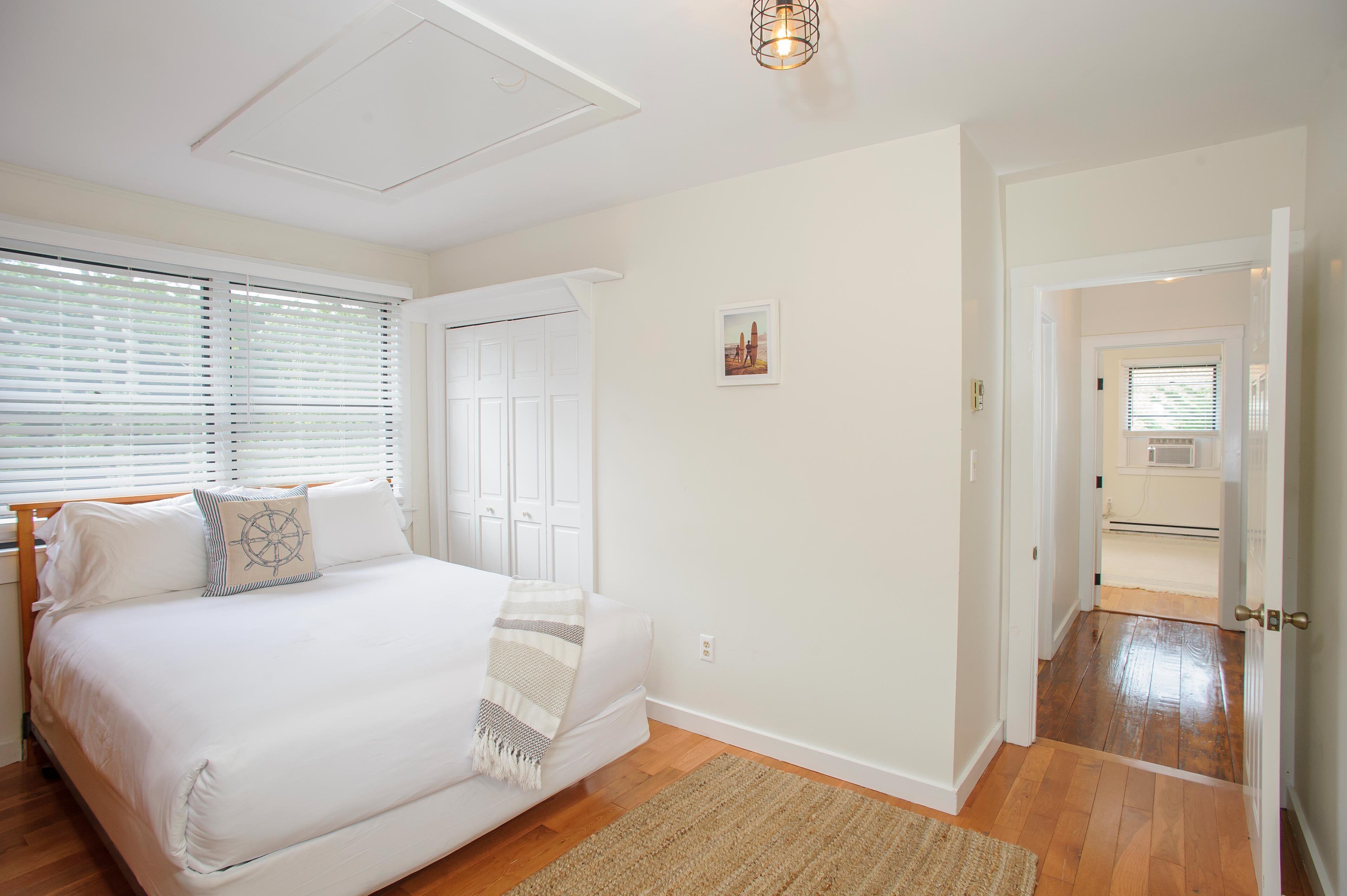A bedroom with a large bed, a wooden floor, a white door, and a hallway leading to another room.