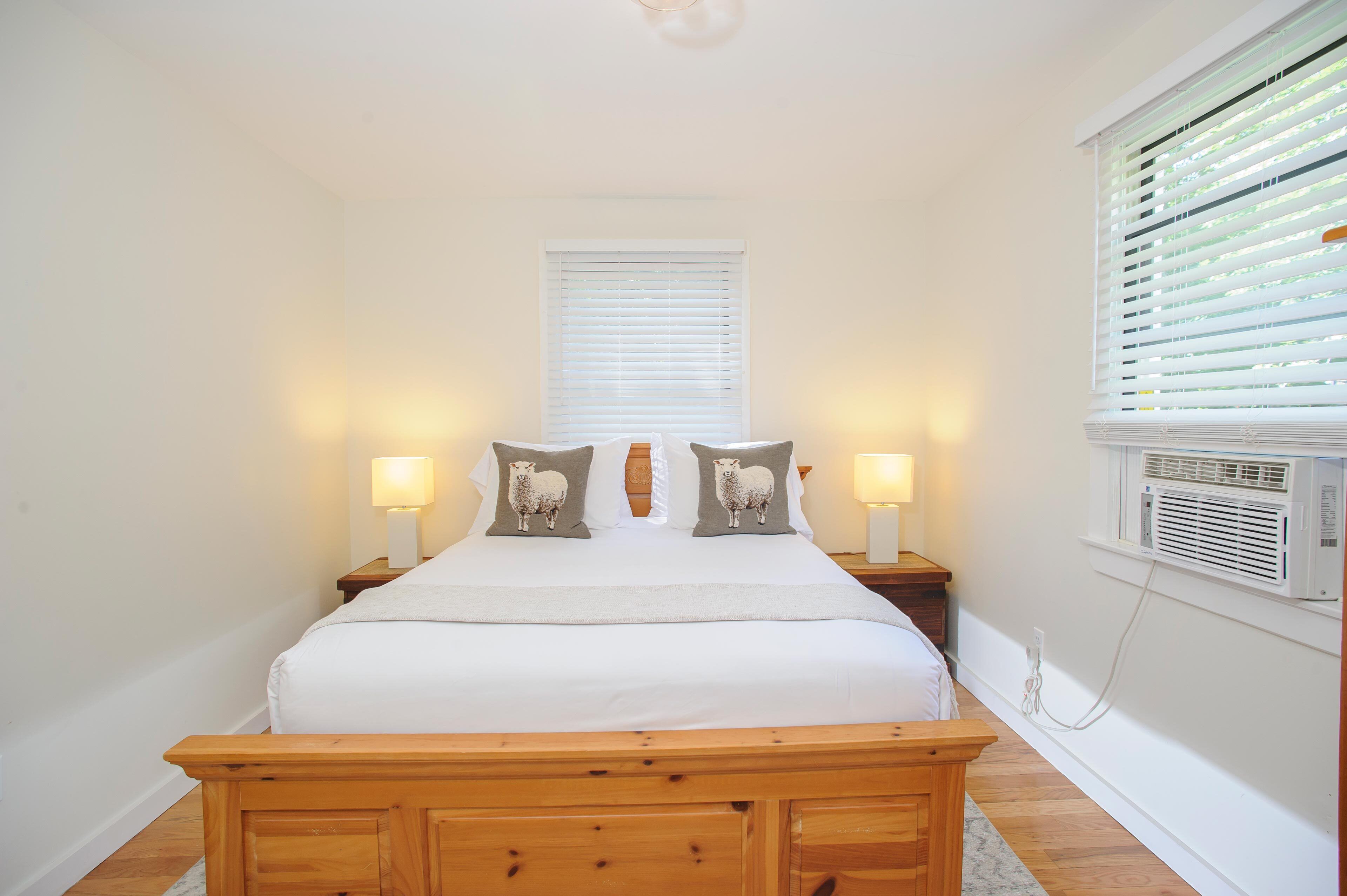 A neatly arranged bedroom with a bed, two bedside lamps, a wooden bench, a window with blinds, and a wall-mounted air conditioning unit.