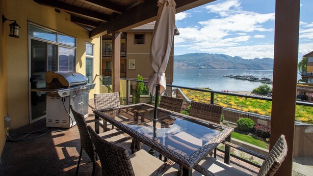 Une terrasse couverte équipée d’un barbecue, de chaises en osier et d’une table en verre, offrant une vue sur un lac où l’on peut apercevoir des bateaux, ainsi que sur des montagnes en arrière-plan.