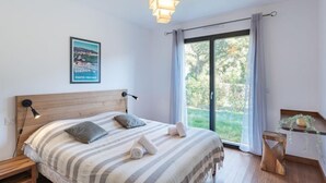 Een slaapkamer met een bed, een nachtkastje, een bedlampje, een houten hoofdbord, een raam met gordijnen en uitzicht op groen buiten.