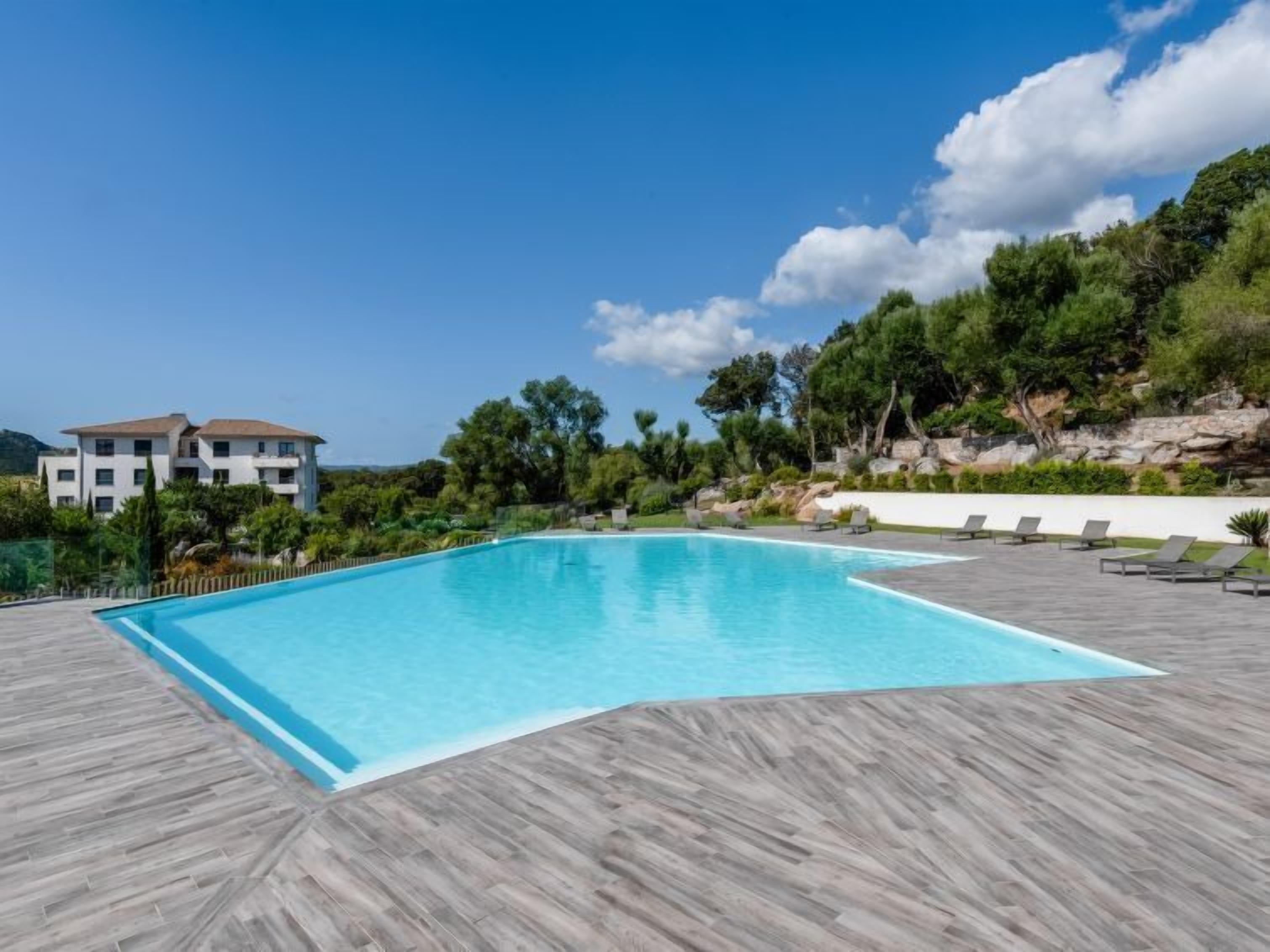 A rectangular swimming pool with a wooden deck, surrounded by lounge chairs and a building in the background.