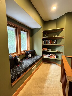 A cozy reading nook with a built-in bench, a bookshelf, and a window with a view of greenery.