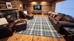 Interior de uma cabana de madeira com lareira, assentos de couro, uma televisão e um tapete xadrez.