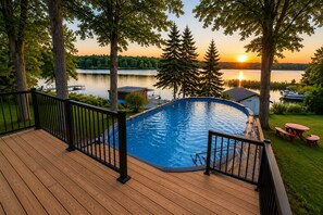 Une piscine dotée d’une terrasse en bois et de rampes noires, entourée d’arbres et offrant une vue sur un lac au coucher du soleil.