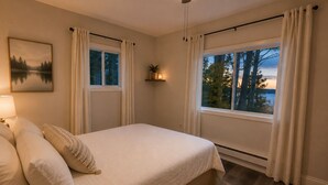 Et soveværelse med en seng, et natbord, en lampe, et vindue med gardiner, en vægmonteret hylde med en plante og et maleri af en sø.