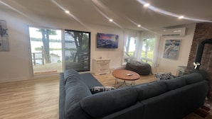 En rummelig stue med en stor sofa, et rundt sofabord og en pejs. Der er en skydedør, der fører til et udendørs område med en træterrasse og udsigt over en sø.