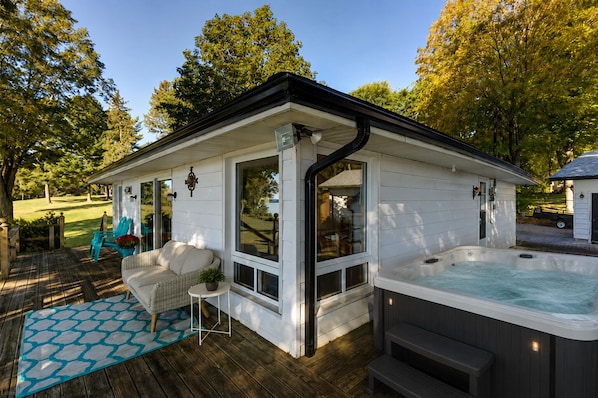 A covered outdoor hot tub area with a wooden deck, a white building with large windows, and a patio with outdoor furniture. - Luxury Waterfront Cottage with Heated Pool, Hot Tub and Dock (Bobcaygeon)