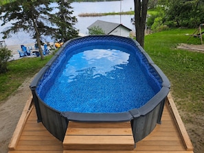 An above-ground swimming pool with a wooden deck, surrounded by a grassy area and trees. - Luxury Waterfront Cottage with Heated Pool, Hot Tub and Dock (Bobcaygeon)