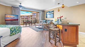 A modern hotel room with a large bed, a TV, a balcony with a view, a dining area, and a kitchen. - Maui Resort Rentals: Honua Kai Hokulani 619 - 6th Floor Studio w/ Full Ocean Views! (Lahaina)