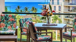 A balcony with wicker chairs and a table, overlooking a beach and ocean with a building in the background. - Maui Resort Rentals: Honua Kai Hokulani 619 - 6th Floor Studio w/ Full Ocean Views! (Lahaina)