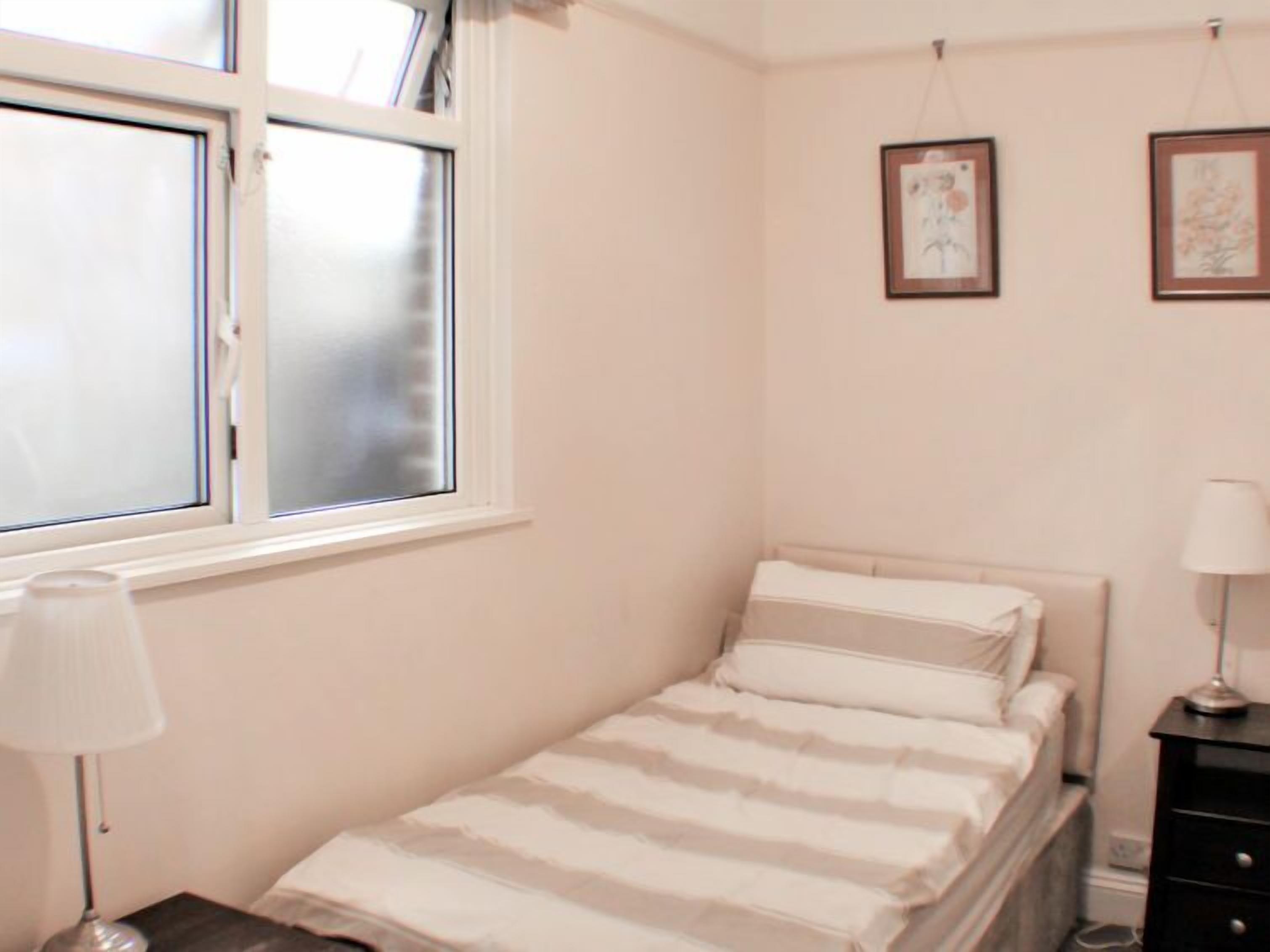 Une petite chambre à coucher, bien rangée, avec un lit simple, une table de chevet, une lampe et une fenêtre à vitre dépolie.