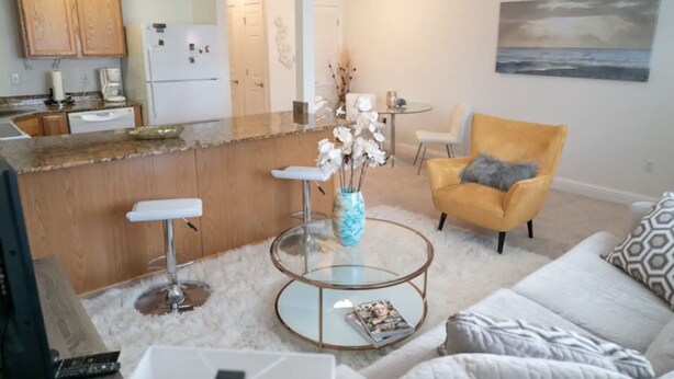 A modern kitchen with wooden cabinets, a white refrigerator, and a dining area with a yellow chair and a white table. - Walk to Beach! King Size Condo! (Biloxi)