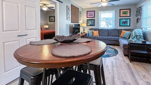 Ein Essbereich mit Holztisch und Barhocker, ein Wohnzimmer mit grauem Sofa und bunten Kissen sowie ein Deckenventilator.