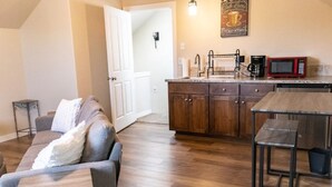Ein gemütliches Wohnzimmer mit grauem Sofa und kleinem Holztisch, eine Küche mit Holzschränken und einer roten Mikrowelle sowie eine weiße Tür zu einem weiteren Raum.