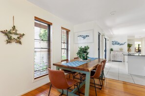 Un comedor con una mesa de madera, cuatro sillas y un mantel individual azul. Hay dos ventanas con persianas, un adorno de estrella en la pared y un cuadro de una ballena.