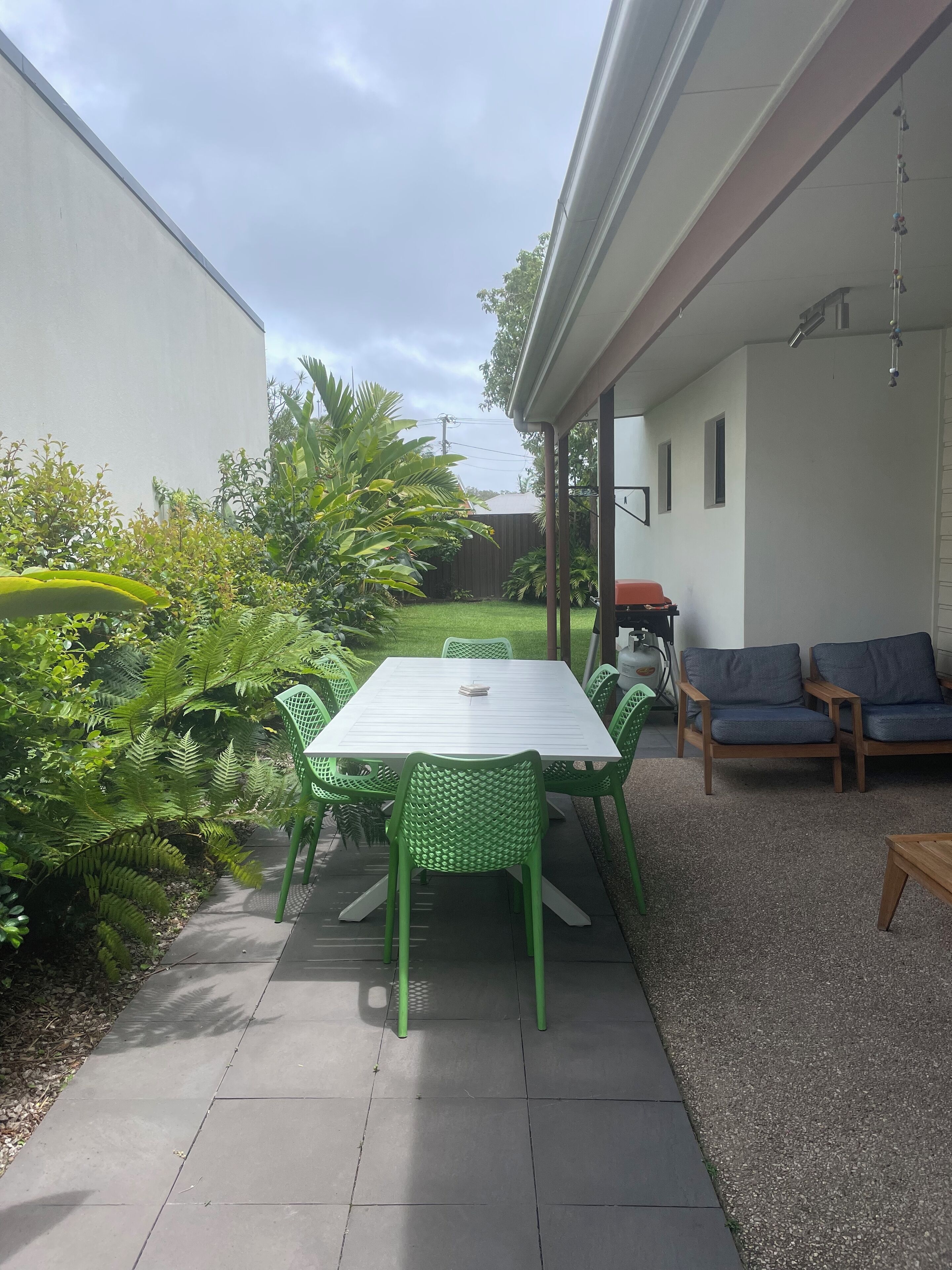 Un patio con una mesa y sillas, rodeado de follaje y una pared blanca.