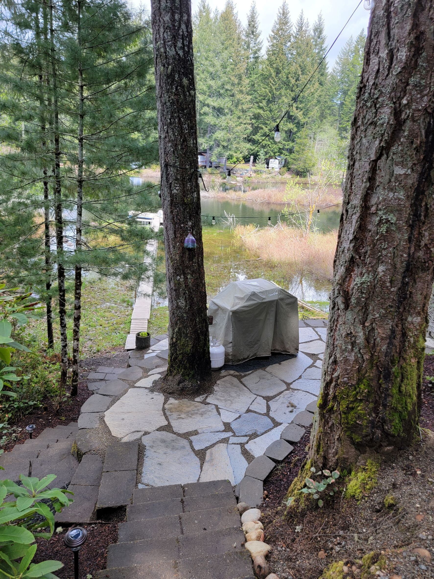 A stone pathway leading to a covered seating area with a table and chairs, surrounded by trees and a body of water.