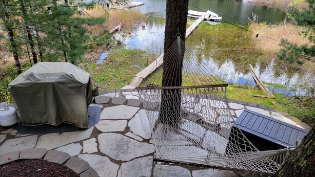 Een terras van steen met een hangmat, een overdekte barbecue en uitzicht op een meer en een steiger.