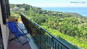 A balcony with a blue chair and a yellow bucket, overlooking a lush green landscape and the sea.