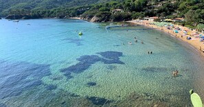 Ein Strand mit klarem, seichten Wasser, einem sandigen Ufer und grünen Hügeln im Hintergrund.