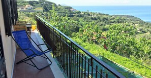 Un balcone con una sedia blu e un secchio giallo, con vista su un paesaggio verde e il mare.