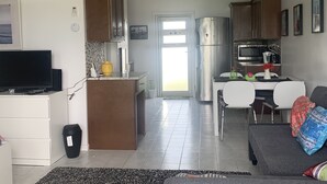 Eine moderne Küche mit Edelstahlgeräten, ein Essbereich mit weißem Tisch und Stühlen sowie ein Wohnzimmer mit einer Couch und einem Fernseher.