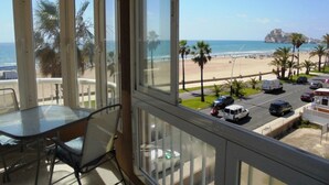 Une pièce avec une table et des chaises en verre, une vue sur une plage bordée de palmiers et un espace de stationnement avec des véhicules.