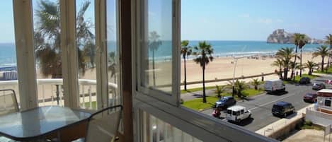 A room with a glass table and chairs, a view of a beach with palm trees, and a parking area with vehicles.