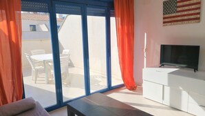 Un salon comprenant un canapé, une table basse, un téléviseur posé sur un meuble blanc et une porte-fenêtre à glissière avec des rideaux orange.