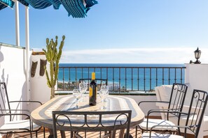 Een ronde tafel met stoelen, een fles en glazen op een terras met uitzicht op zee.