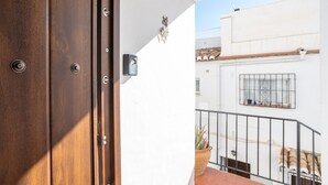 Une porte en bois avec une poignée en métal, un petit luminaire fixé au mur, et une vue sur un bâtiment blanc doté d’une fenêtre et d’une plante en pot.