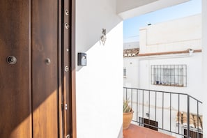 A wooden door with a metal handle, a small wall-mounted light, and a view of a white building with a window and a potted plant.