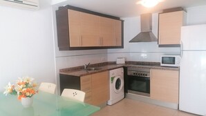 A compact kitchen with a washing machine, microwave, stove, and a small dining area with a glass table and chairs. - Miralcielo 2ºA- LEK (Peñiscola)