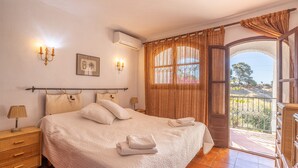 Un dormitorio con una cama grande, una mesita de noche, una lámpara, aire acondicionado instalado en la pared, un cuadro y un balcón con vistas.