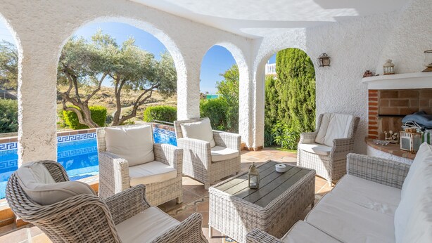 Un espacio exterior techado con mobiliario de mimbre, una chimenea y vistas a una piscina y árboles.