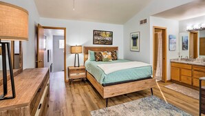 A bedroom with a wooden bed, a nightstand, a lamp, a ceiling fan, a bathroom with a sink and mirror, and a door leading to another room. - Cozy Vail Valley Townhome - Walk to Riverwalk! (Edwards)