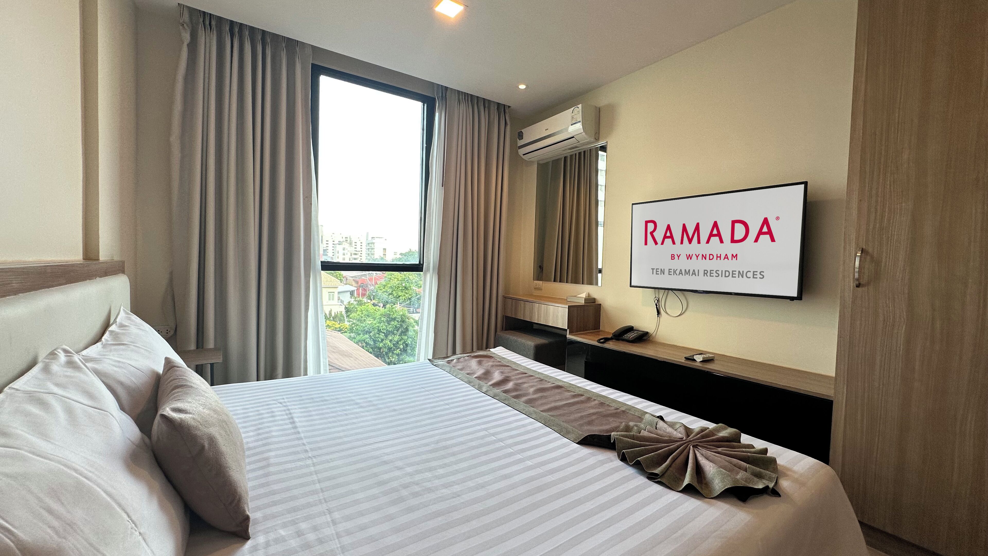 A hotel room with a large bed, a desk with a phone, a TV, and a view of the city through the window.