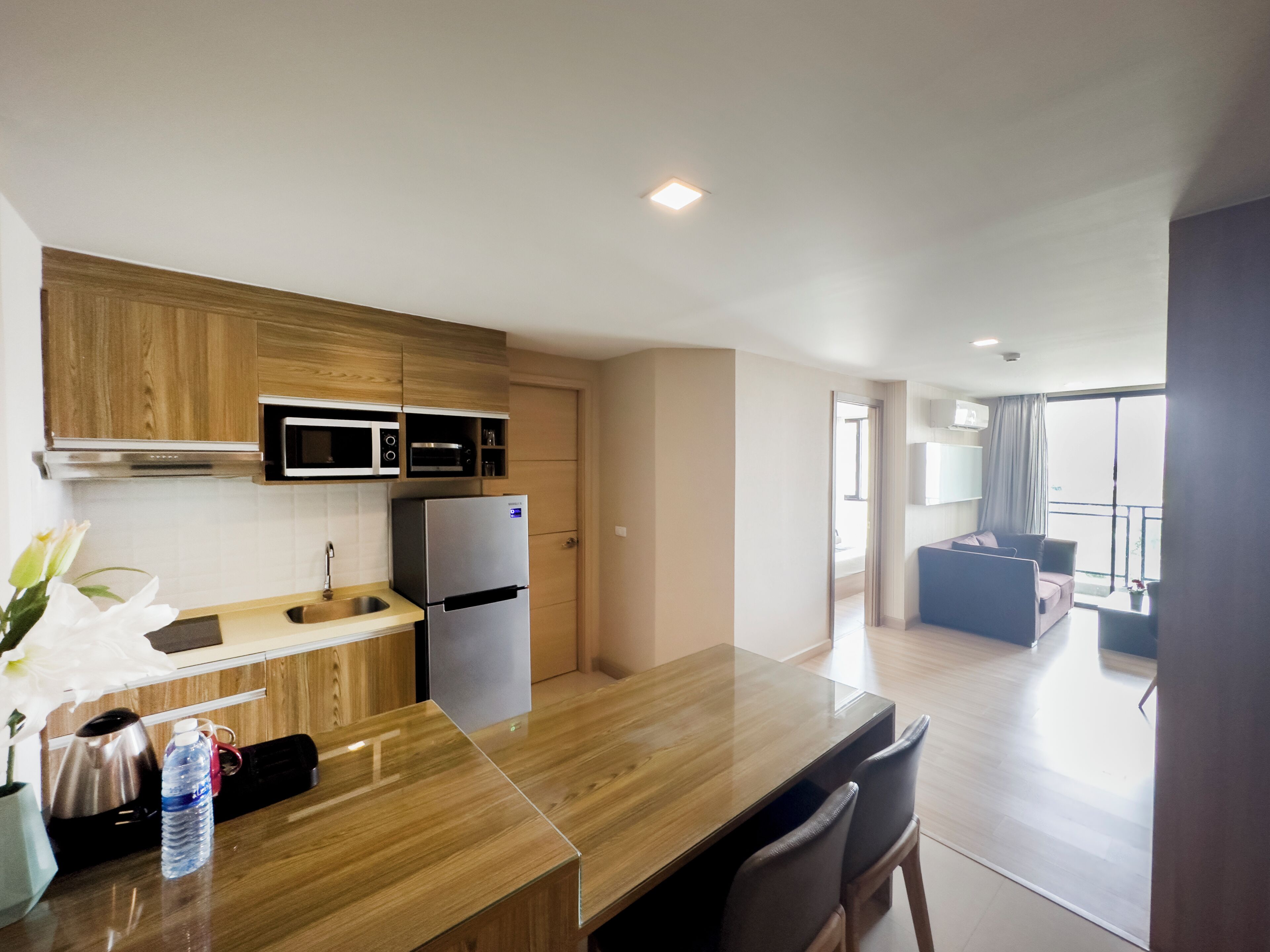 Una cocina moderna con armarios de madera, un refrigerador de acero inoxidable y un microondas. Hay un comedor con una mesa y sillas de madera, y vistas a un balcón con un sofá.
