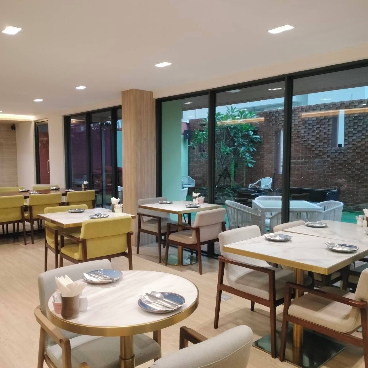 A restaurant with round and rectangular tables, yellow chairs, and a view of a brick wall and greenery outside.
