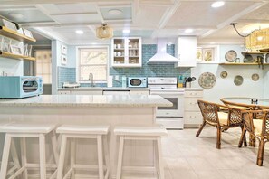 A modern kitchen with a central island, white cabinetry, and a dining area with wicker chairs. - Walk to Ocean: Home w/ Fire Pit in Atlantic Beach (Atlantic Beach)
