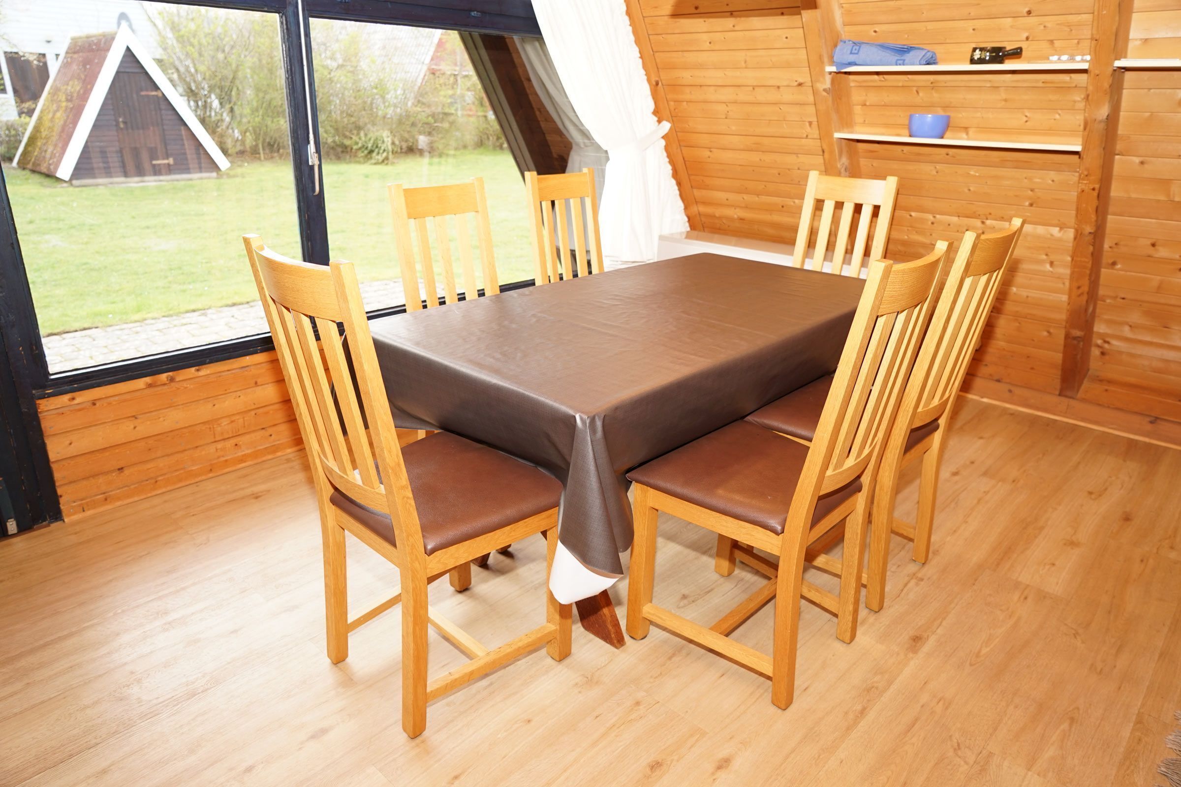 Een houten eettafel met zes stoelen, uitzicht op een grasveld en een houten gebouw buiten, plus een plank met spullen aan de muur.