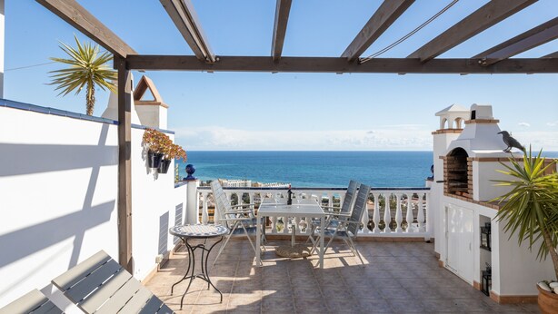 Um alpendre coberto com vista para o mar, uma mesa com cadeiras e uma pérgula de madeira.