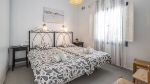 Uma cama de casal com roupa de cama estampada, uma mesa de cabeceira com abajur, um candeeiro pendente, um quadro na parede e uma janela com cortinas.
