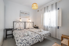 Una cama doble con ropa de cama estampada, una mesita de noche con lámpara, una lámpara colgante, un cuadro en la pared y una ventana con cortinas.