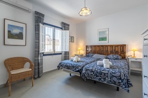 A bedroom with a double bed, two bedside tables with lamps, a wicker chair, and a window with curtains.