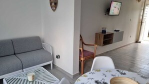 Een woonkamer met een grijze bank, een witte salontafel, een rieten bijzettafeltje, een witte stoel en een kleine eettafel met een bloemendessin.
